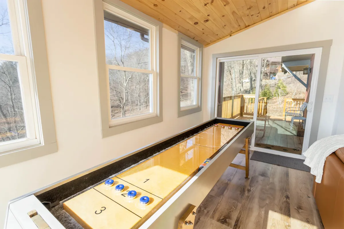 The photo features a shuffleboard table positioned beneath a wooden ceiling, flanked by expansive windows that showcase natural light and glimpses of the outdoors. The space feels open and playful, perfect for socializing and enjoying mountain scenery.