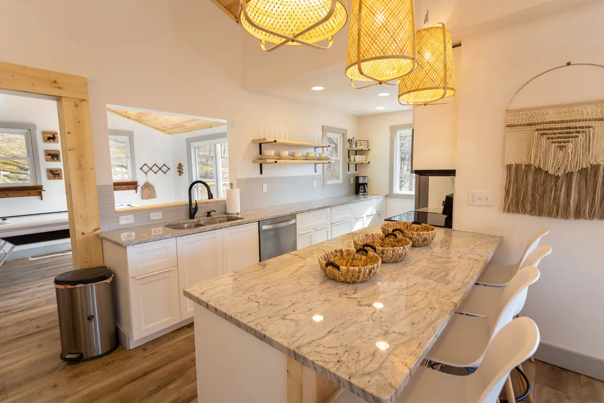 A spacious, modern kitchen featuring a large granite-topped island with seating for four. Sleek stainless steel appliances line the walls, open shelves display decorative pieces, and elegant pendant lights hang overhead. The overall scene radiates a sense of openness and invites group cooking and socializing.