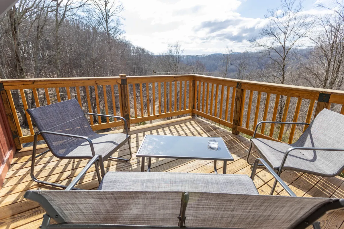 A serene deck seating area looks out onto panoramic mountain views, with a table and glass suggesting casual drinks or al fresco meals. The setup evokes peaceful mornings or social afternoons, with the mountains providing a dramatic backdrop for conversation and relaxation.