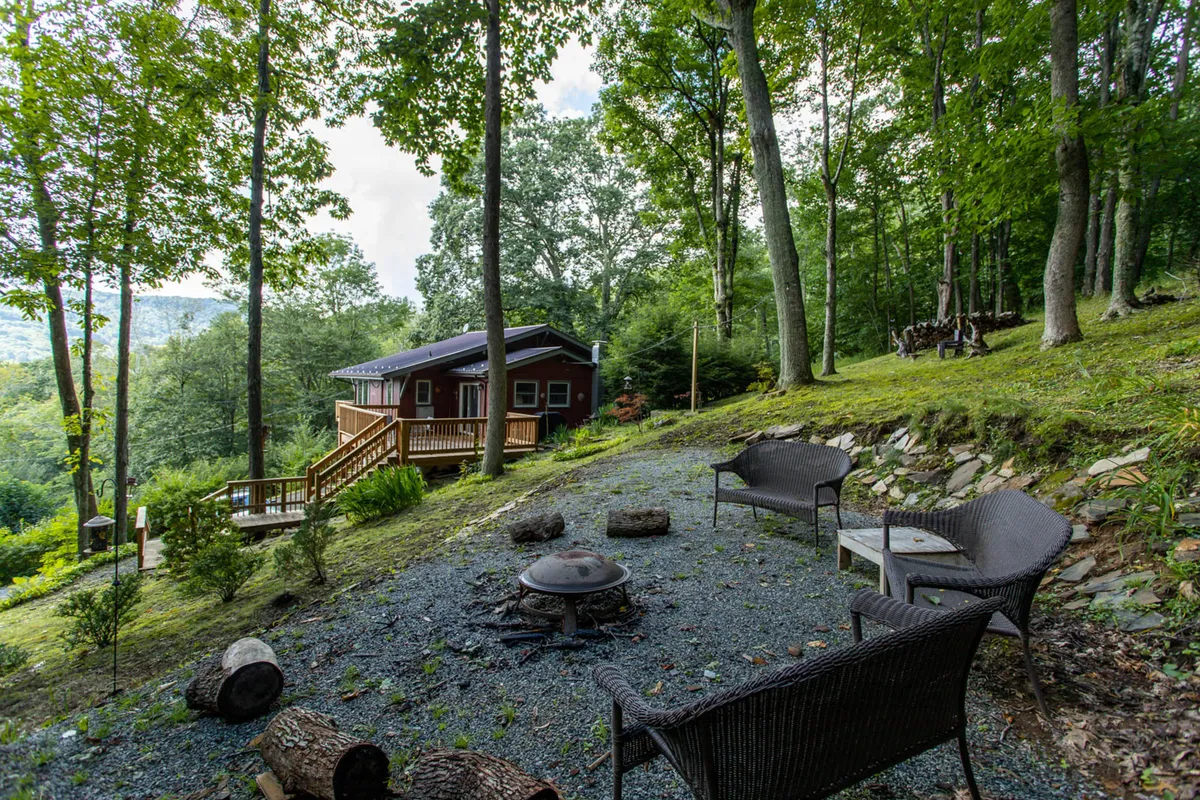 This image showcases a cozy outdoor fire pit area surrounded by comfortable seating and set against a backdrop of trees and the inviting cabin. The scene is designed for relaxed campfire gatherings, storytelling, stargazing, and sharing laughter under the open sky.