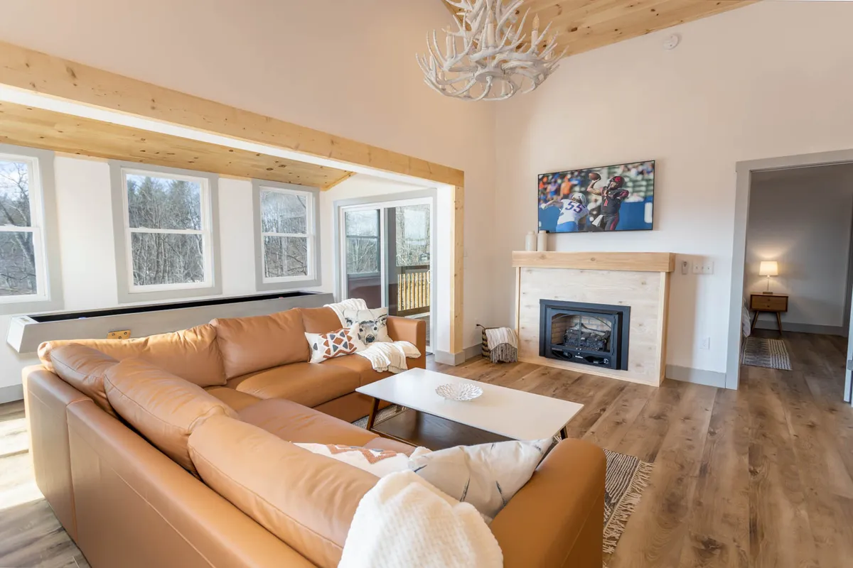 The image captures the great room’s inviting atmosphere with a large leather sofa arranged around a glowing gas log fireplace. Above, an antler chandelier gleams, adding rustic charm. Artwork accents the walls, and the space is designed for relaxing, entertaining, and movie nights, all beneath soaring ceilings.