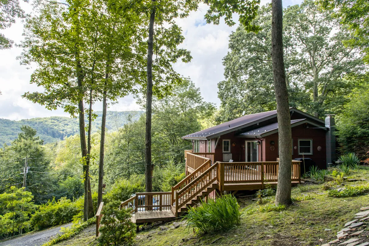 The image showcases the inviting front of the red chalet, nestled amidst lush trees on a hillside, with a spacious wooden deck offering panoramic mountain views. The early morning light or late evening glow casts a serene ambiance, making the deck an ideal spot for relaxation, socializing, or admiring nature. The setting promises privacy, tranquility, and a strong connection to the picturesque surroundings.