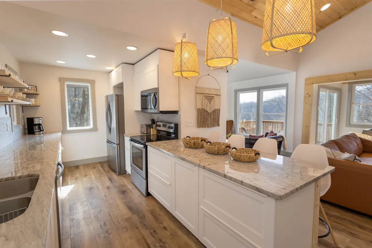 The kitchen is shown from a slightly different angle, with the island, pendant lights, and stainless steel appliances all visible. The granite countertop is prominent, and the mood feels warm and inviting for family cooking and coffee rituals.