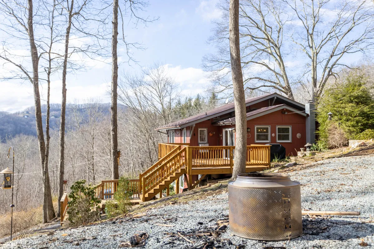 A cozy fire pit set on the wooden deck, with the house’s welcoming facade and natural landscape in the background. The scene invites gatherings at dusk, with hints of warmth and camaraderie against the backdrop of the wilderness.