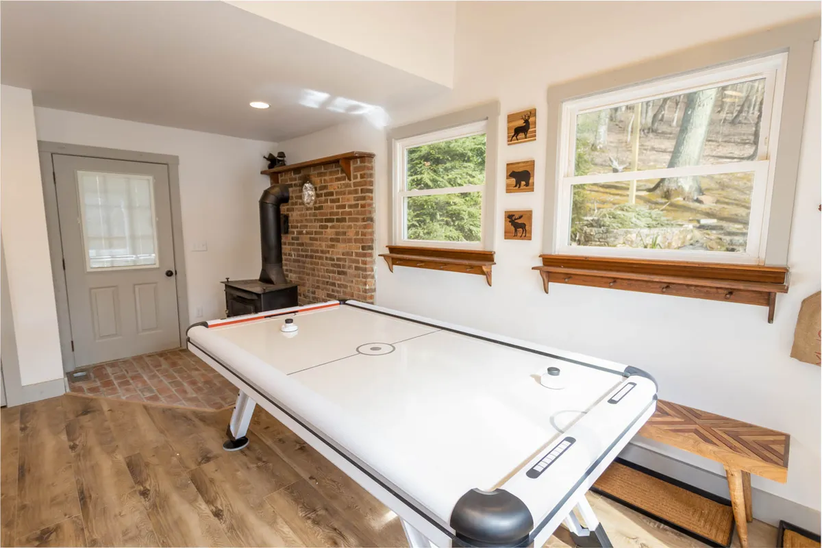 This fun, energetic image spotlights the air hockey table in a cozy nook by a brick fireplace. Large windows behind the table frame scenic views of the outdoors, allowing natural light to fill the room and offering glimpses of the mountains beyond.