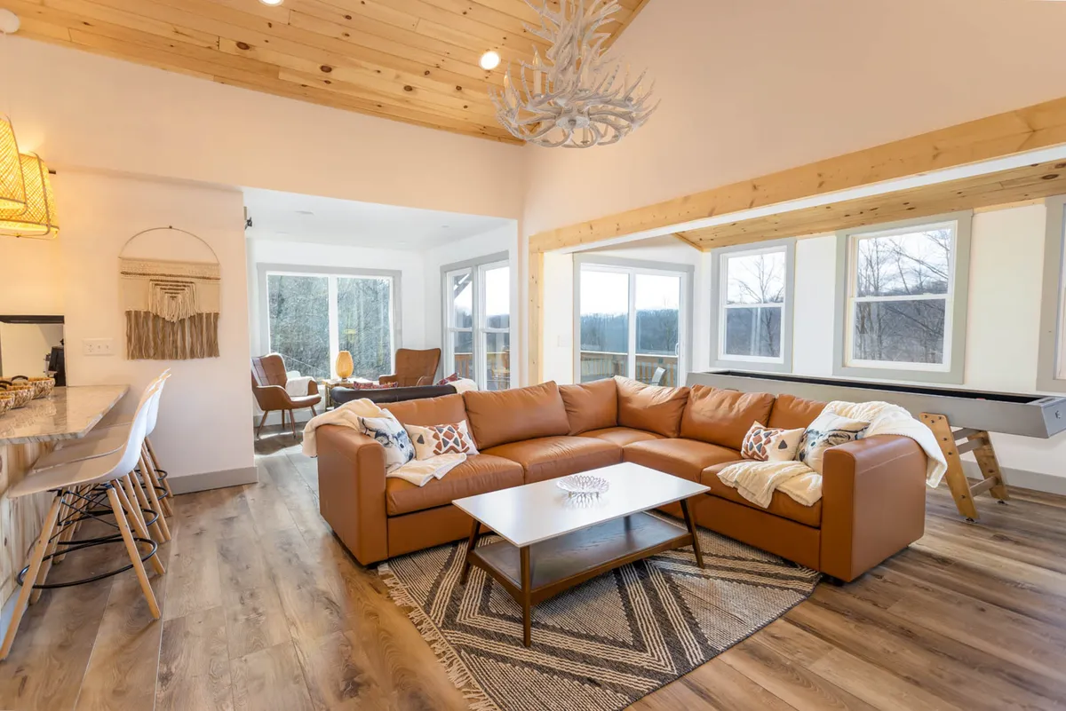 This wide-angle view unites the living area, dining space, and kitchen. A leather sectional sofa faces large windows revealing mountain scenery, with a chandelier overhead and a wooden dining table nearby. The open-concept layout keeps everyone connected.