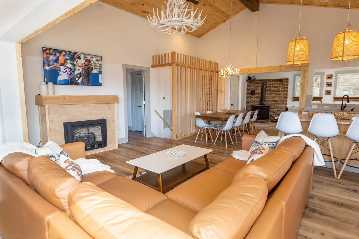 A spacious and inviting living room with a leather sofa, crackling fireplace, dining table, and television. The layout encourages family and friends to gather, with the sofa offering additional sleeping space for guests.